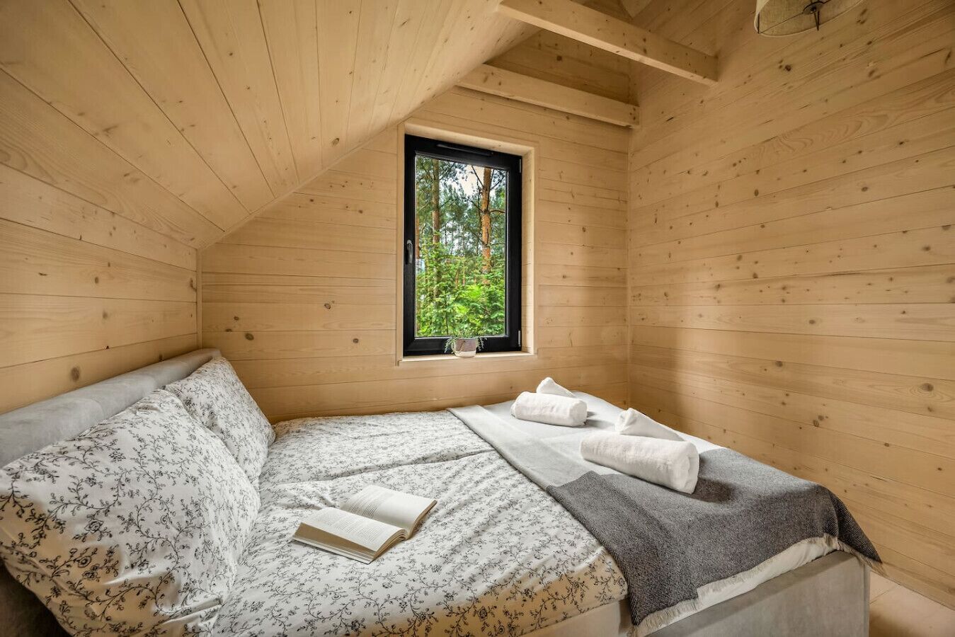 Un dormitori de cabana de fusta amb un llit, una finestra que mostra arbres i un llibre sobre el llit.
