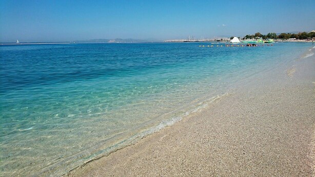 Una playa con aguas poco profundas y transparentes, una orilla arenosa y un paisaje urbano distante.
