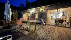 An outdoor wooden deck with a dining table and chairs, a stone wall, and a glass door leading to an interior kitchen area. - Pink Granite Coast in Brittany: Comfortable Home with Wi-Fi (Trébeurden)
