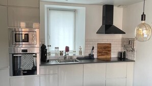 A modern kitchen with a built-in oven, microwave, and sink. There is a window with a view of the outside, and a dining area with a table and chairs. - Pink Granite Coast in Brittany: Comfortable Home with Wi-Fi (Trébeurden)