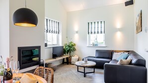 Ein modernes Wohnzimmer mit einer Couch, einem Fernseher, einem Esstisch mit Hut und Champagner sowie einer großen Pendelleuchte.