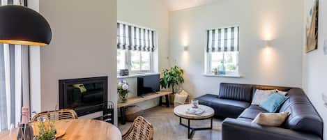 A modern living room with a black leather sofa, a wooden dining table with a tray, a TV, and a large window with striped blinds.