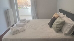 A neatly made bed with pillows and folded towels, a radiator, and a window with a view of the outside. - Neos Kosmos Cozy Retreat - Cental Comfort (Athens)