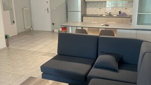 A modern living room with a sectional sofa, a wooden coffee table, and a kitchen area with light wood cabinets and a refrigerator. - Neos Kosmos Cozy Retreat - Cental Comfort (Athens)