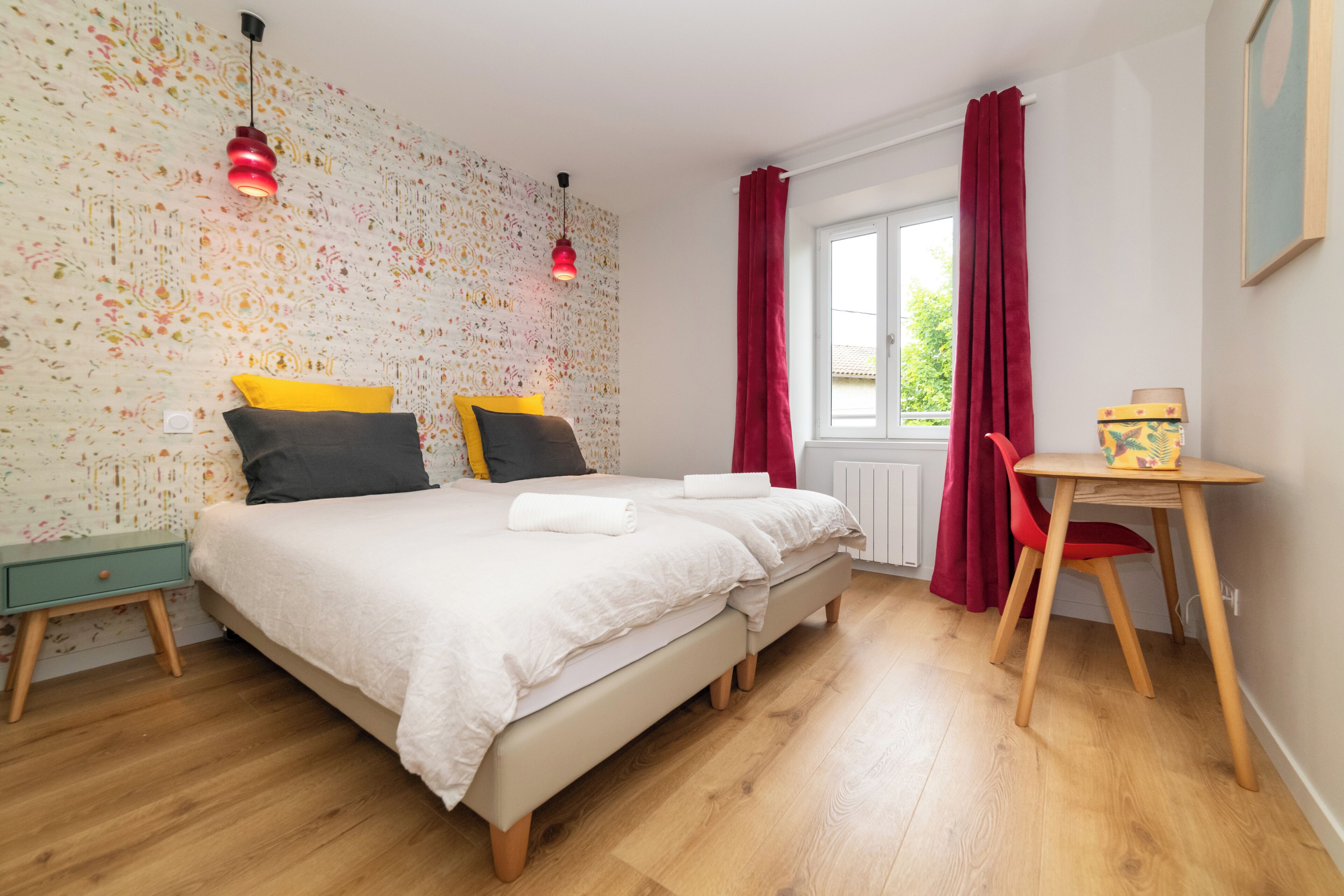 Een slaapkamer met een bed, nachtkastjes, een raam met gordijnen en een klein tafeltje met een stoel.