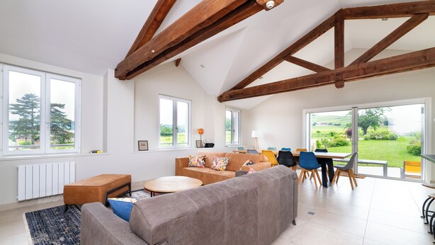 A spacious living area with a large sofa, a dining table with chairs, and a view of the outdoors through large windows. - L'échappée Beaujolaise (Juliénas)