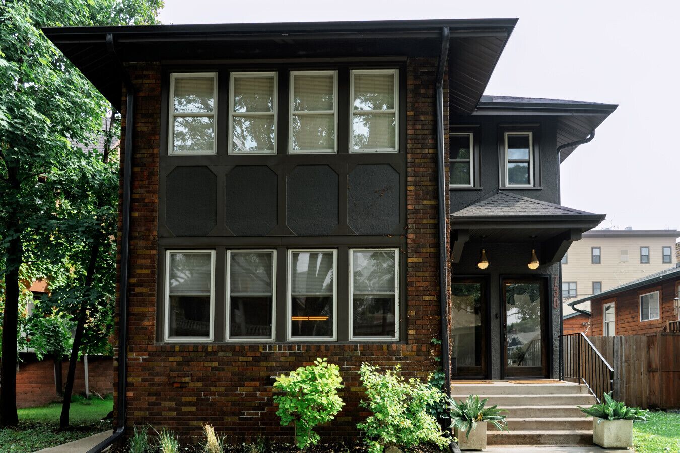 Une maison à deux étages avec une façade en briques, de grandes fenêtres et un porche à l’avant avec des marches.