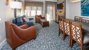 A hotel room with a blue patterned carpet, a brown leather sofa, a blue armchair, a wooden dining table with chairs, and a stone fireplace.