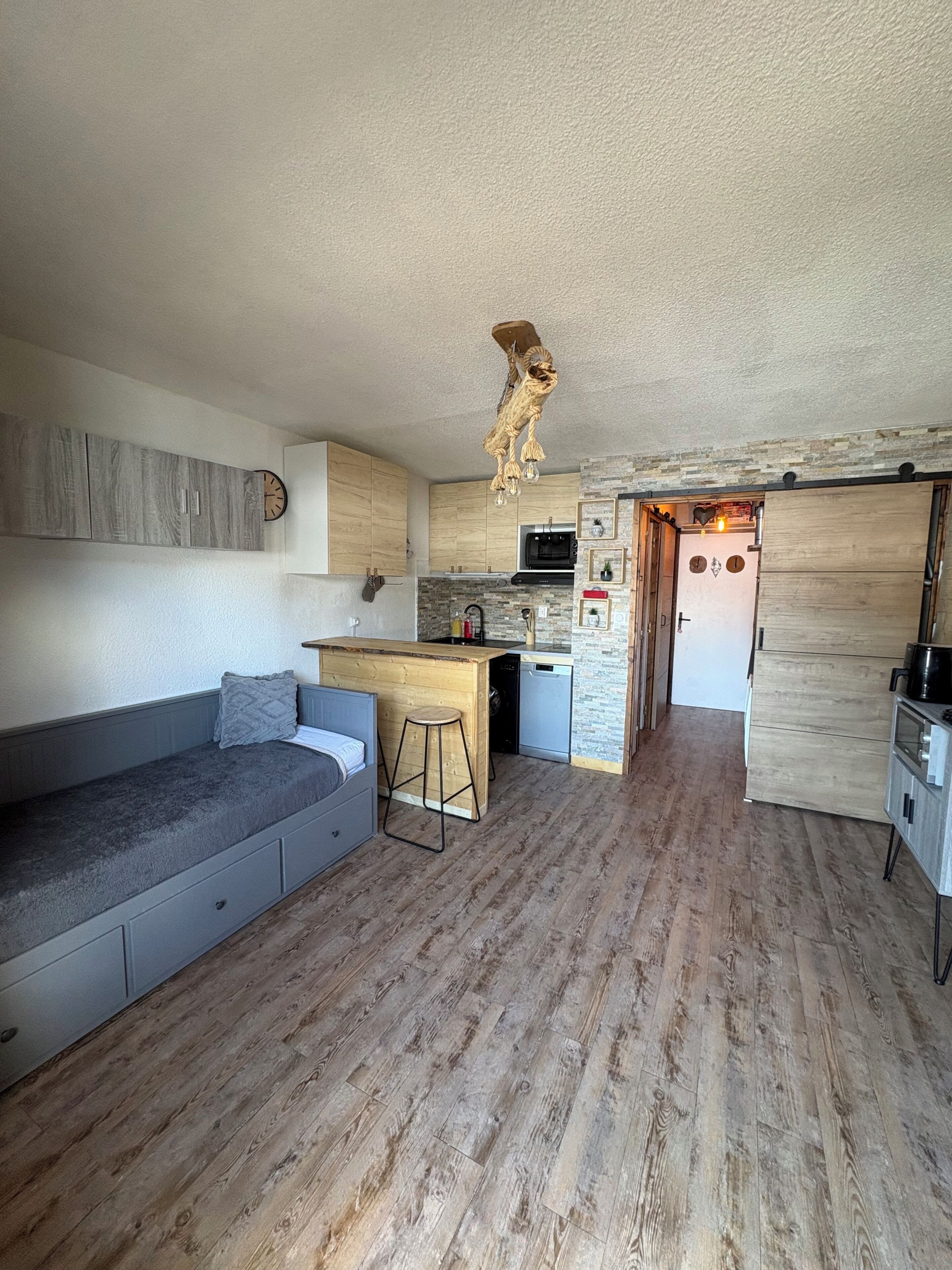 A compact living space with a kitchenette, a wooden floor, a grey sofa, and a unique ceiling light fixture.