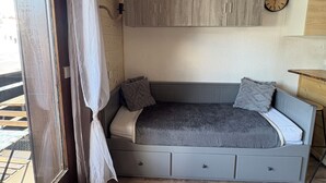 Un dormitorio pequeño con cama, piso de madera, ventana con cortinas, un reloj en la pared y un mueble de madera.