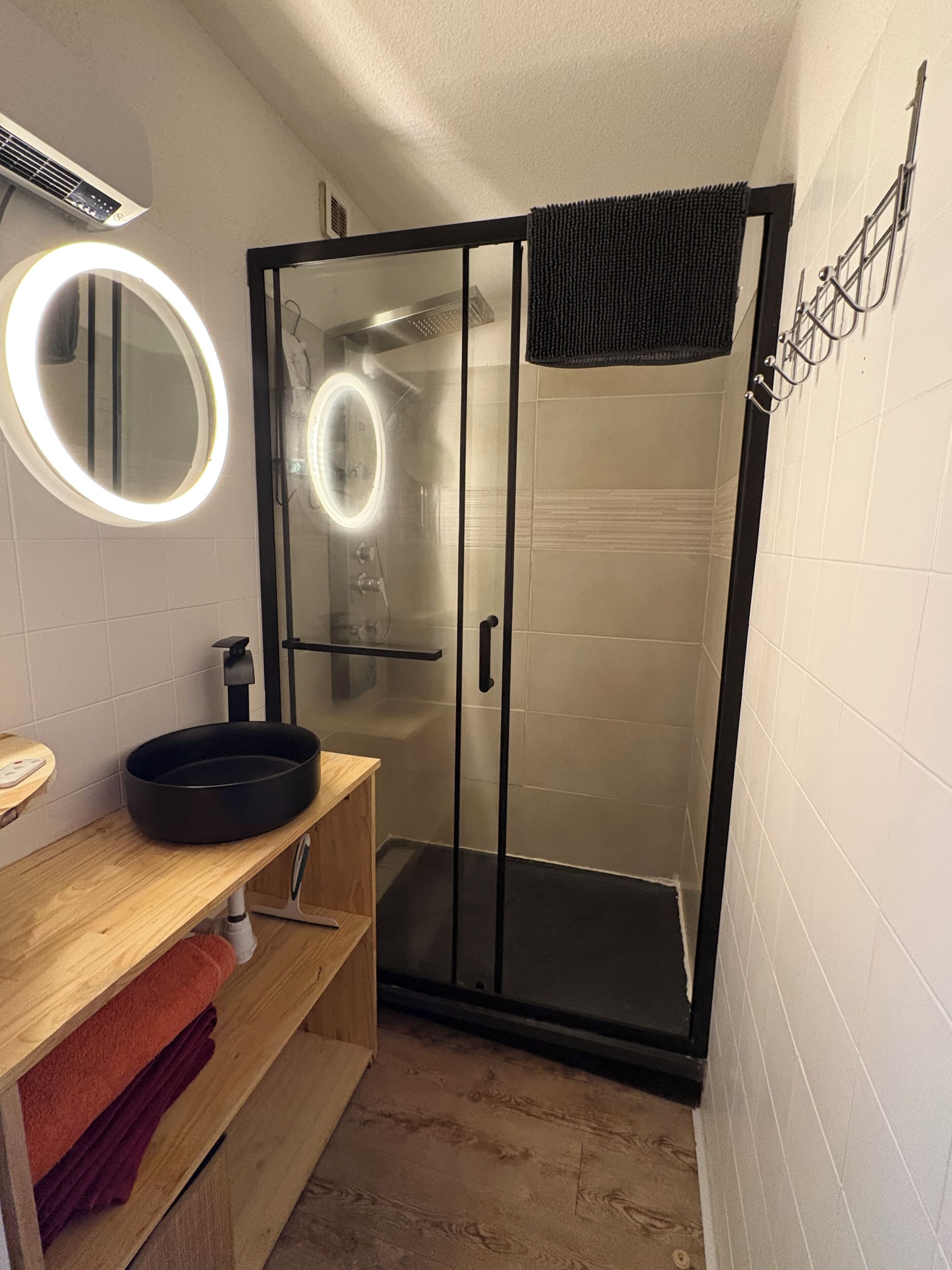 Une salle de bain avec une douche à parois de verre, un miroir rond avec un éclairage intégré et un meuble-lavabo en bois avec un vasque.