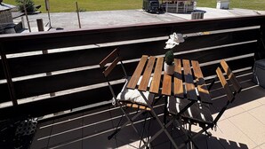 Un patio exterior con una mesa y sillas de madera, un paraguas rojo y vistas a un paisaje verde con árboles y una montaña al fondo.