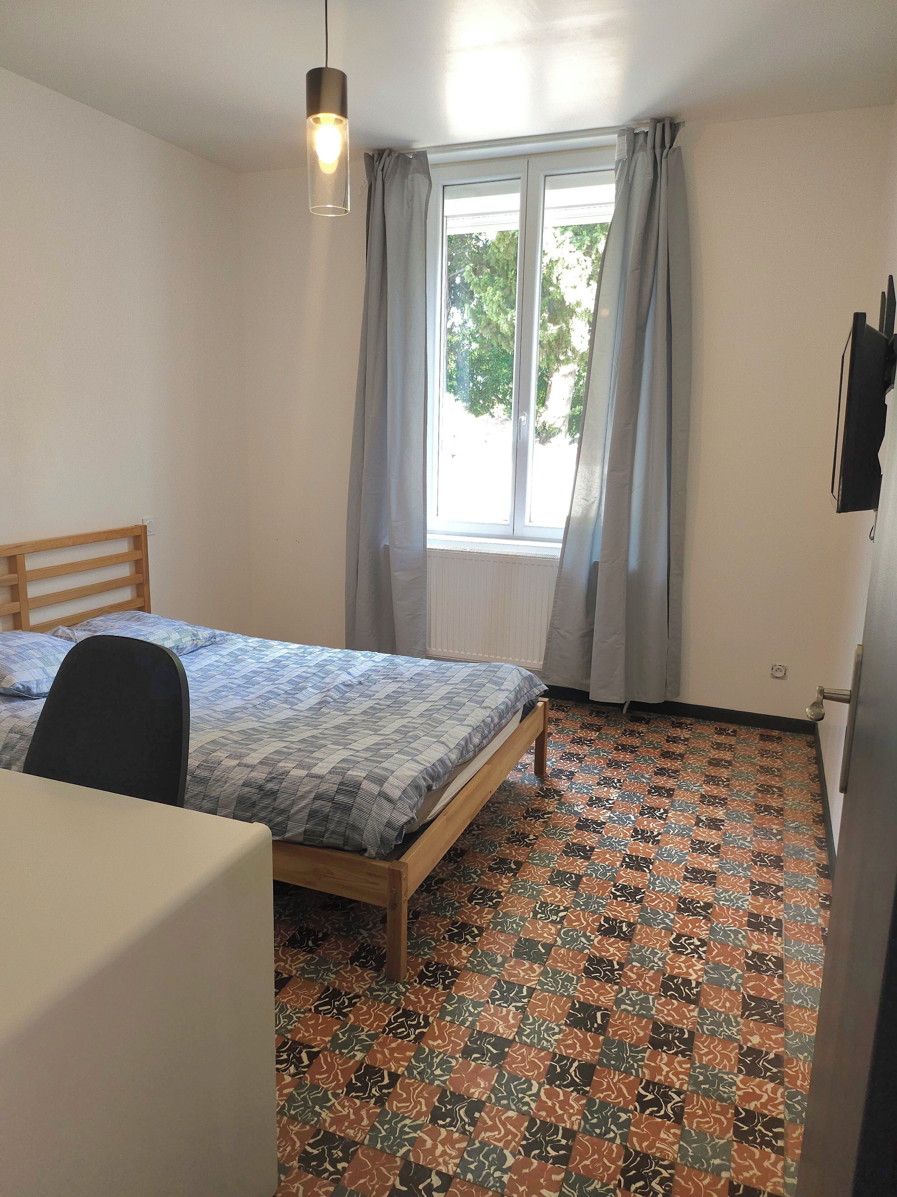 A bedroom with a bed, a chair, a desk, a window with curtains, and a patterned floor.