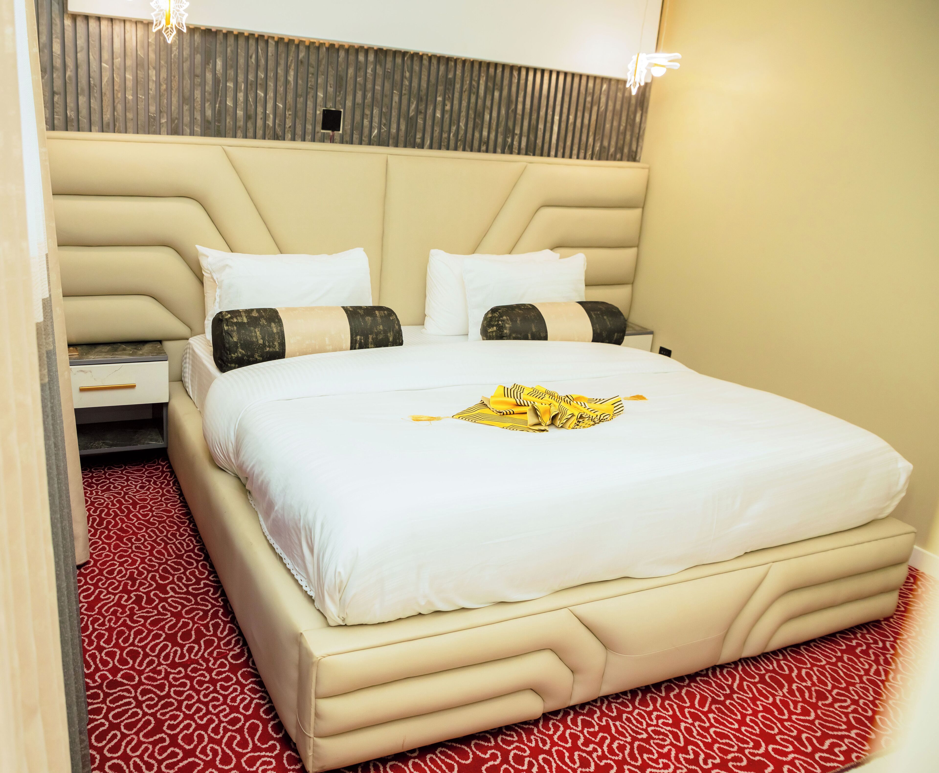 A hotel room with a large bed, a headboard, a nightstand, and a red patterned carpet.