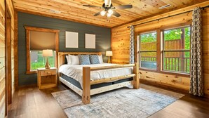 Un dormitorio de cabaña de madera con una cama, dos lámparas de noche, un ventilador de techo y ventanas grandes con cortinas.