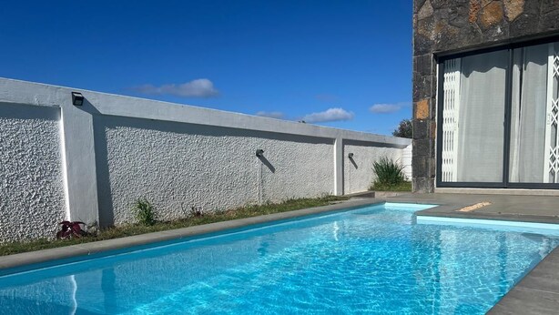 Ein rechteckiger Swimmingpool mit klarem, blauem Wasser, umgeben von einer Steinmauer und einem modernen Gebäude mit großen Fenstern.