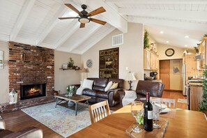 Ein gemütliches Wohnzimmer mit Kamin, Ledersofas, einem Esstisch mit Weingläsern und einer Küche im Hintergrund.
