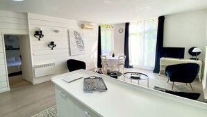 A modern living room with a white sofa, a dining table with chairs, a TV, and a large window with curtains. - T3 curiste rental 600m from the spa (Eugénie-les-Bains)