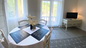 A dining area with a round table set for breakfast, lunch, and dinner, featuring a clock on the wall and a TV on a small table. - T3 curiste rental 600m from the spa (Eugénie-les-Bains)