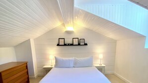 A bedroom with a sloped ceiling, a bed with white linens, two bedside lamps, a wooden headboard, and a small shelf with framed pictures. - T3 curiste rental 600m from the spa (Eugénie-les-Bains)