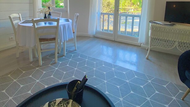 A room with a dining table and chairs, a TV on a white cabinet, and large windows with curtains. - T3 curiste rental 600m from the spa (Eugénie-les-Bains)