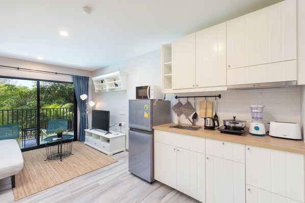 A modern kitchen with white cabinets, a stainless steel refrigerator, a blender, and a toaster. There is a dining area with a table and chairs, and a balcony with a view of greenery.