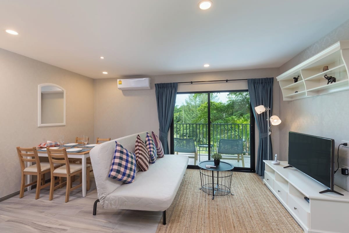 A modern living room with a sofa, dining table, chairs, a TV, and a balcony with a view of greenery.