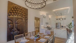 A modern dining area with a wooden table set for six, a large Buddha painting on the wall, and a kitchen in the background. - NEW Spacious 5-bedroom house with Views, Spa, Fire pit, Golf & Hammocks (Sedona)