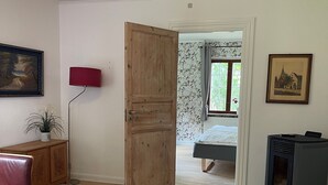 Een kamer met een houten tafel, een rode lamp, een klein kastje, een vloerkleed, een bed, een raam met gordijnen, en een verwarming.