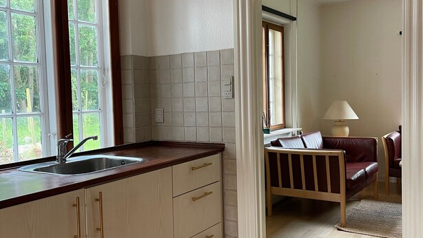 Een keuken met houten kasten, een spoelbak en een raam. Een doorgang leidt naar een slaapkamer met een bed en nachtkastje.