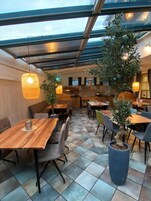 Um restaurante com decoração moderna, com mesas e cadeiras de madeira, plantas em vasos e teto de vidro.