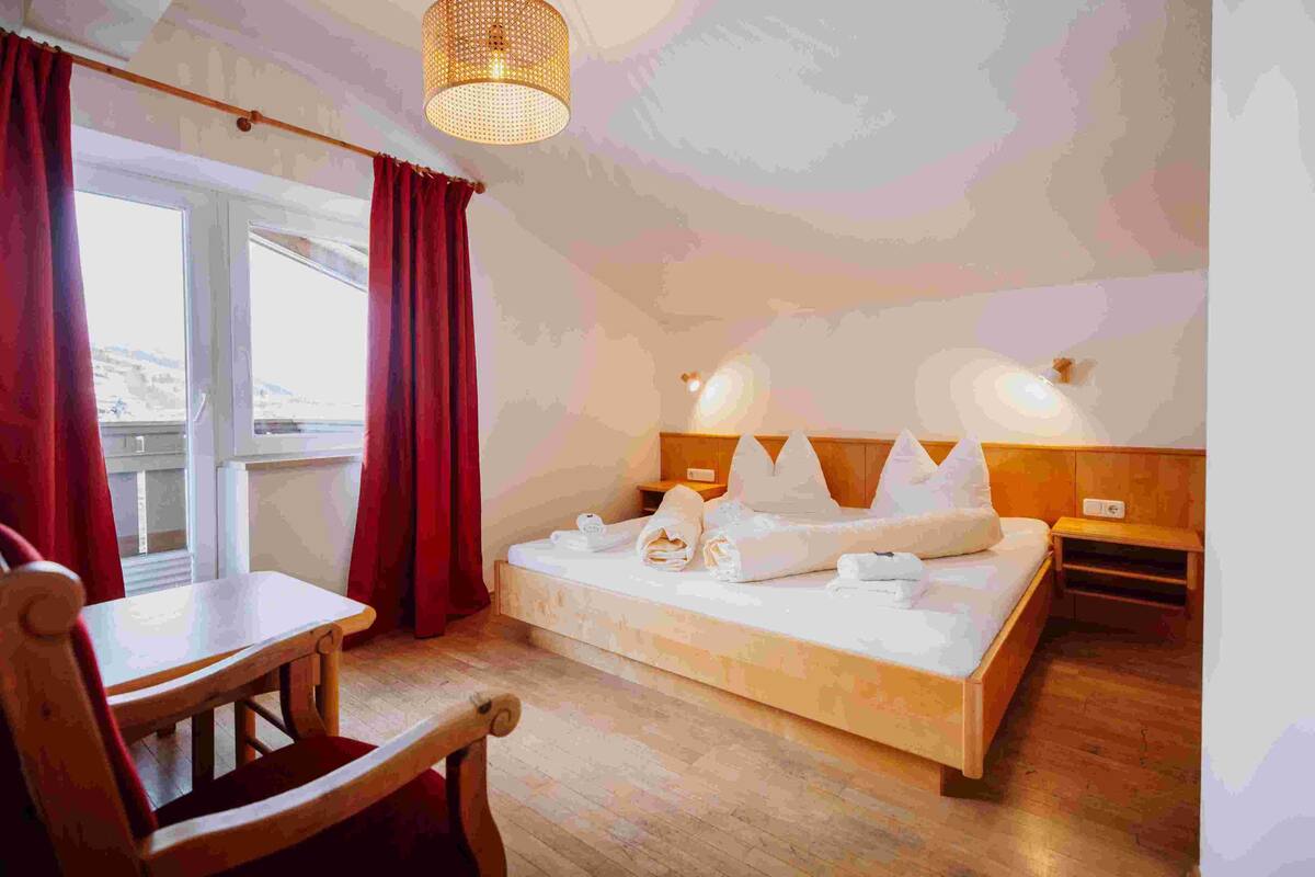 A bedroom with a wooden bed, a wooden chair, a table, and a window with red curtains.