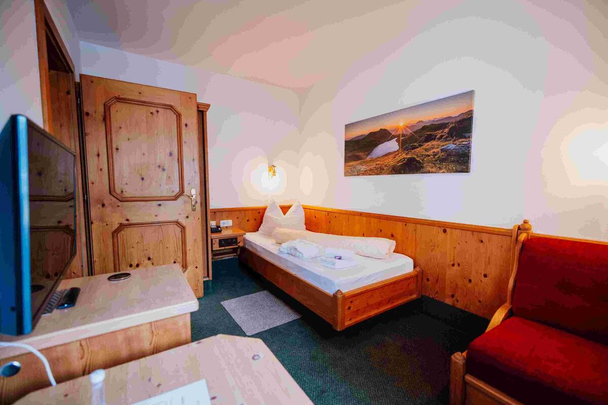 A room with a wooden bed, a red armchair, a desk with a computer, and a wall-mounted TV.