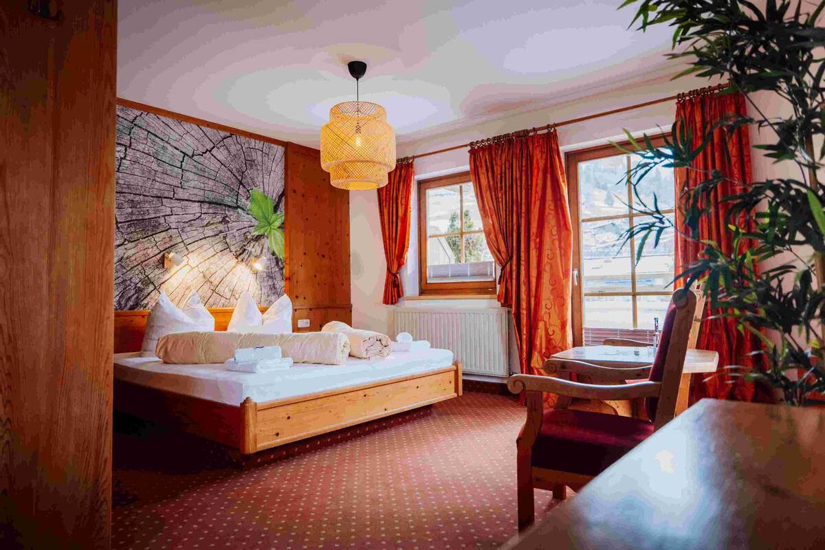 A bedroom with a wooden bed, a desk, a chair, a large window with curtains, and a hanging light fixture.