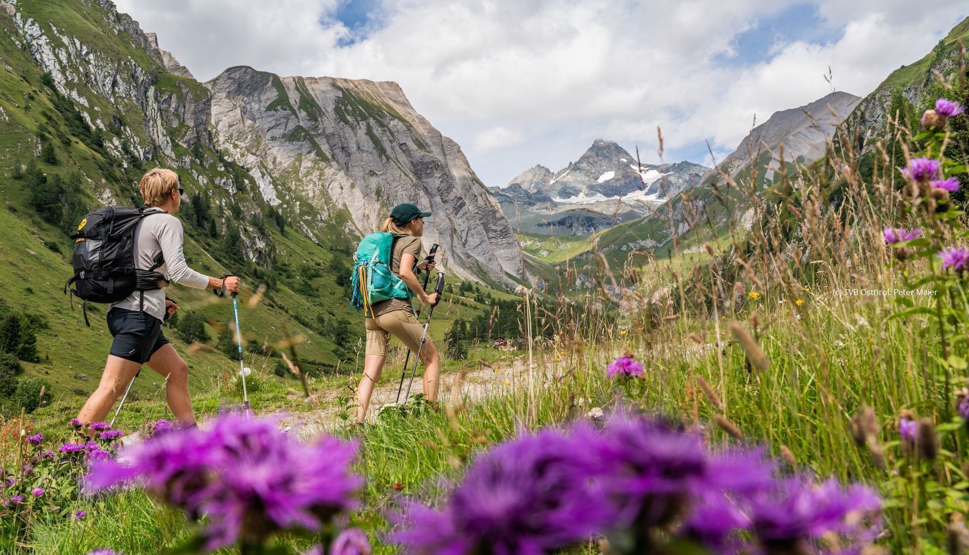 Two hikers on a mountain trail with rocky mountains in the background.
