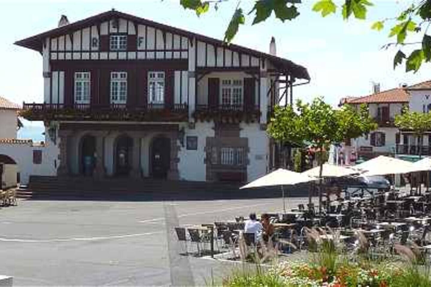 Un bâtiment à deux étages doté d’un balcon, d’une entrée centrale avec des marches et d’un patio aménagé avec des tables et des parasols.