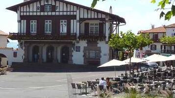 Un bâtiment à deux étages doté d’un balcon, d’une entrée centrale avec des marches et d’un patio aménagé avec des tables et des parasols.