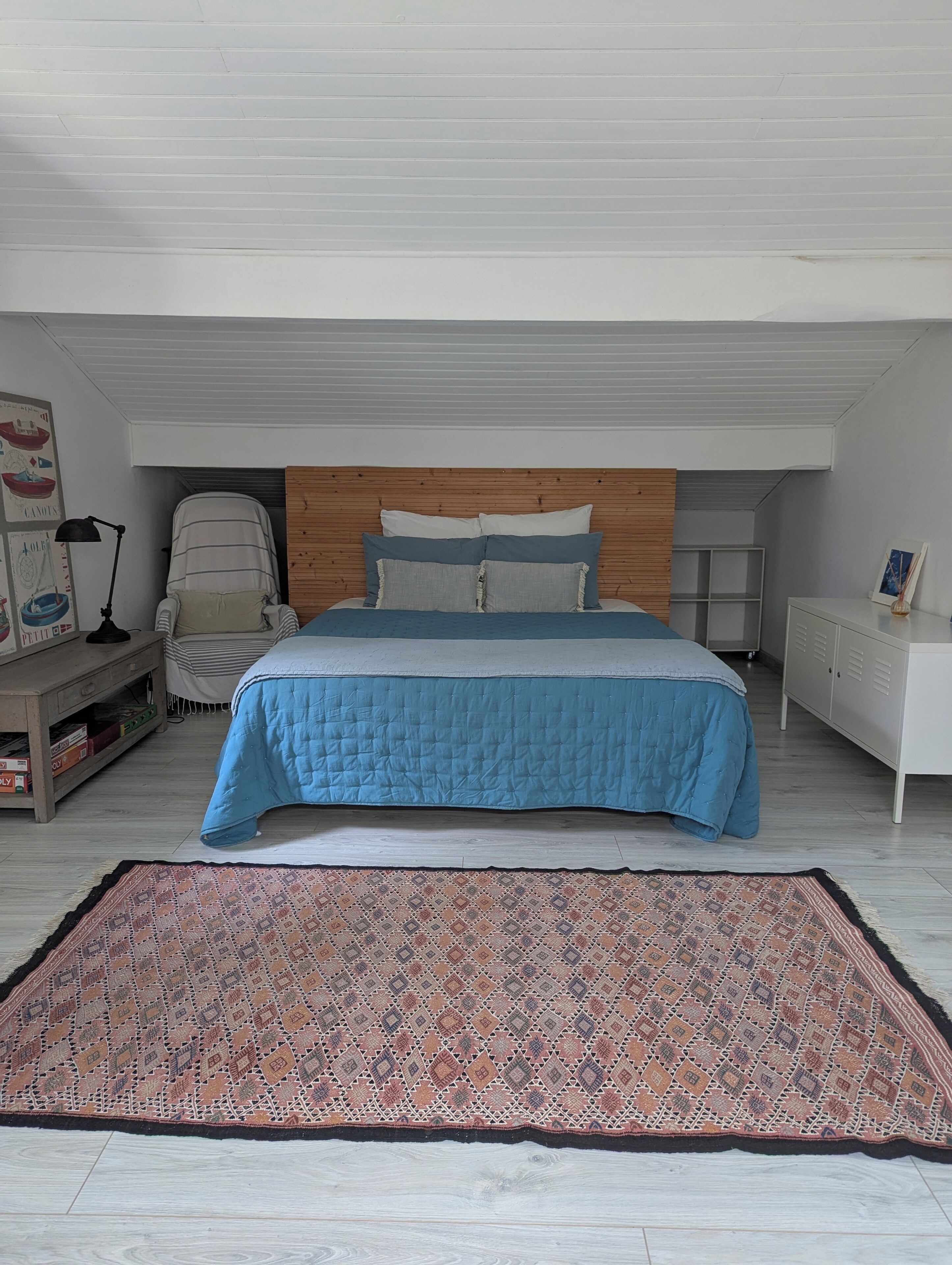 A bedroom with a bed, a wooden headboard, a bedside table, a chair, a rug, and a wall with artwork.