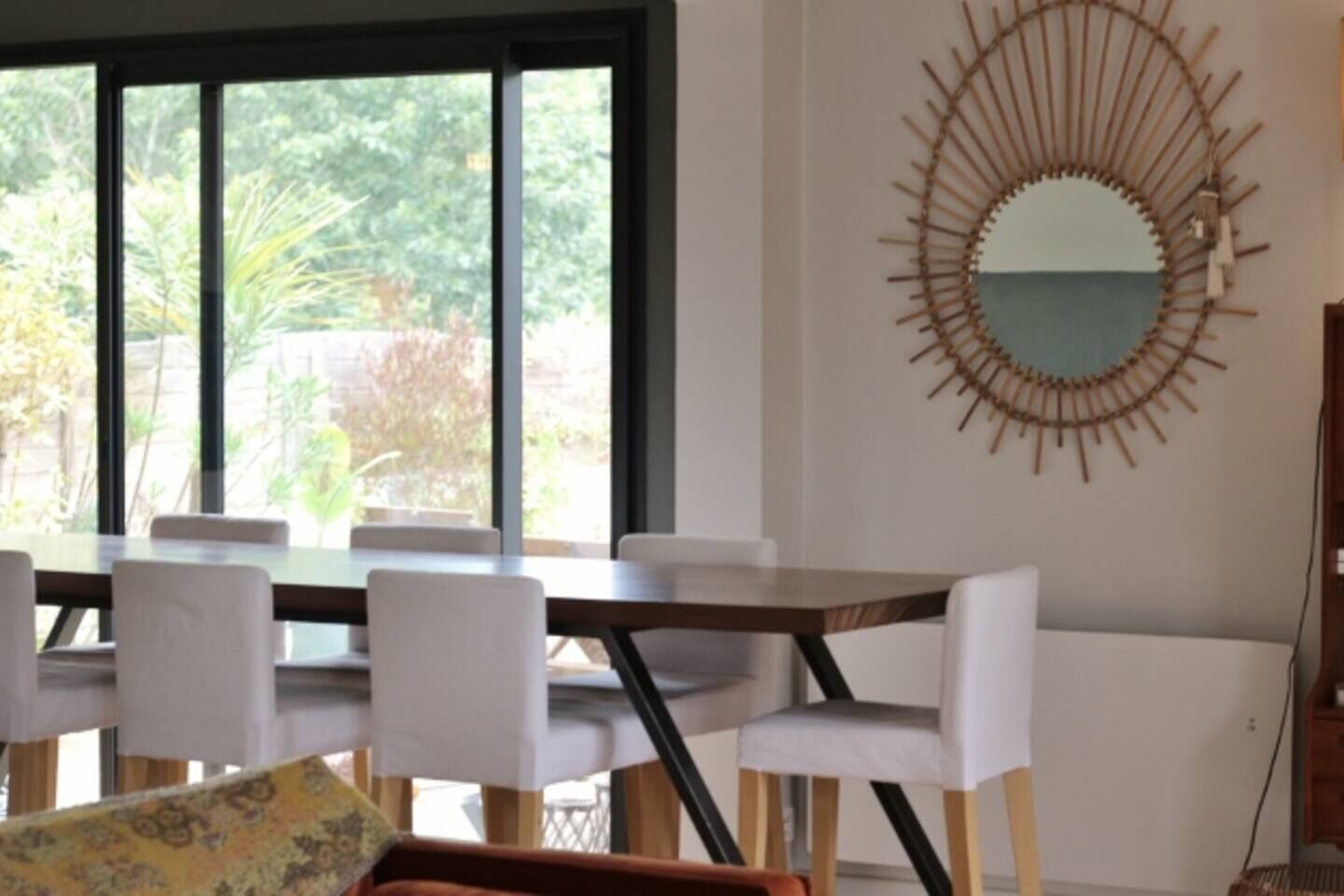 A dining area with a large table and chairs, a decorative wall mirror, and a view of greenery outside.