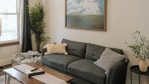 A living room with a grey sofa, a coffee table, a patterned rug, a painting of clouds, and a plant. - Modern 2 Bedroom Downtown Apartment (Milwaukee)