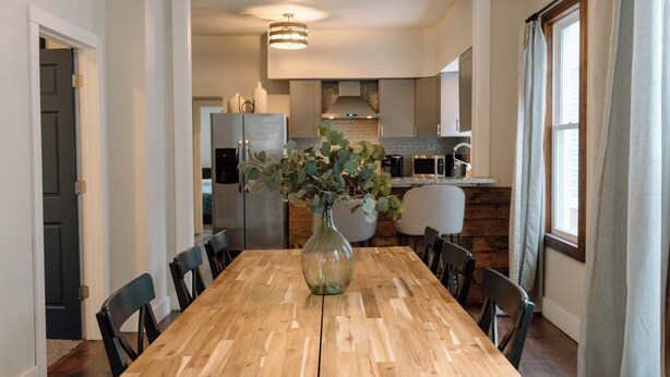 A dining area with a wooden table, chairs, and a vase with greenery. A kitchen is visible in the background with modern appliances. - Modern 2 Bedroom Downtown Apartment (Milwaukee)