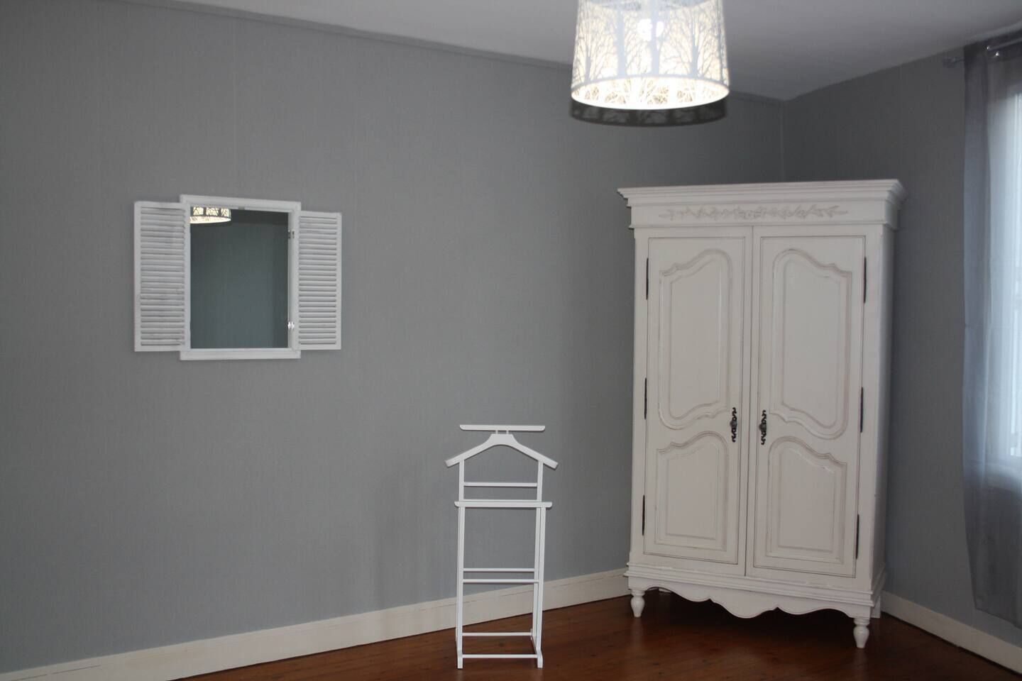 Une pièce aux murs gris, avec une armoire blanche, un portant à vêtements blanc et une fenêtre dotée de volets blancs.