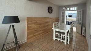 A dining area with a wooden accent wall, a dining table with chairs, a lamp, and a kitchen in the background. - Gîte L'Elixir (Fécamp)