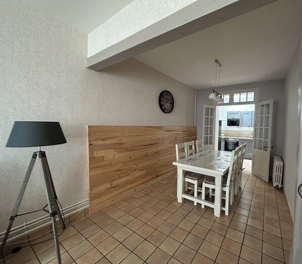 A dining area with a wooden accent wall, a dining table with chairs, a lamp, and a kitchen in the background. - Gîte L'Elixir (Fécamp)