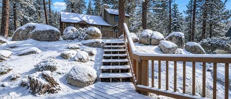 A wooden deck leading to a cabin surrounded by snow-covered rocks and trees.