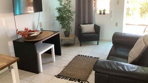 Un salon comprenant un téléviseur, une petite table ornée d’un vase décoratif, un canapé, un fauteuil, une plante en pot et une fenêtre avec des rideaux.
