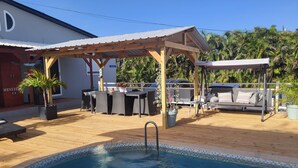 Un espace piscine doté d’une terrasse en bois, d’un coin salon couvert et d’un bâtiment sur lequel est apposée une enseigne indiquant « MINIBIKE ».