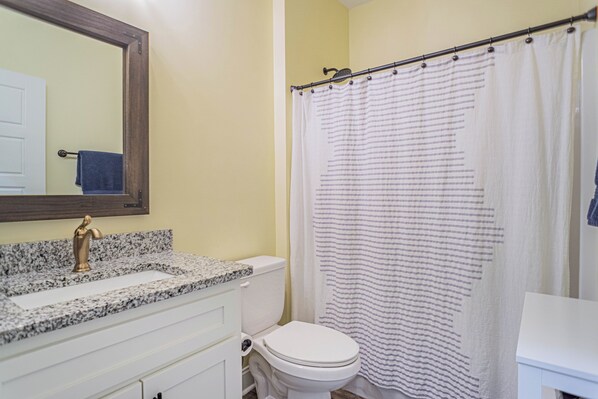 A bathroom with a granite countertop, a white toilet, a shower curtain, and a mirror.