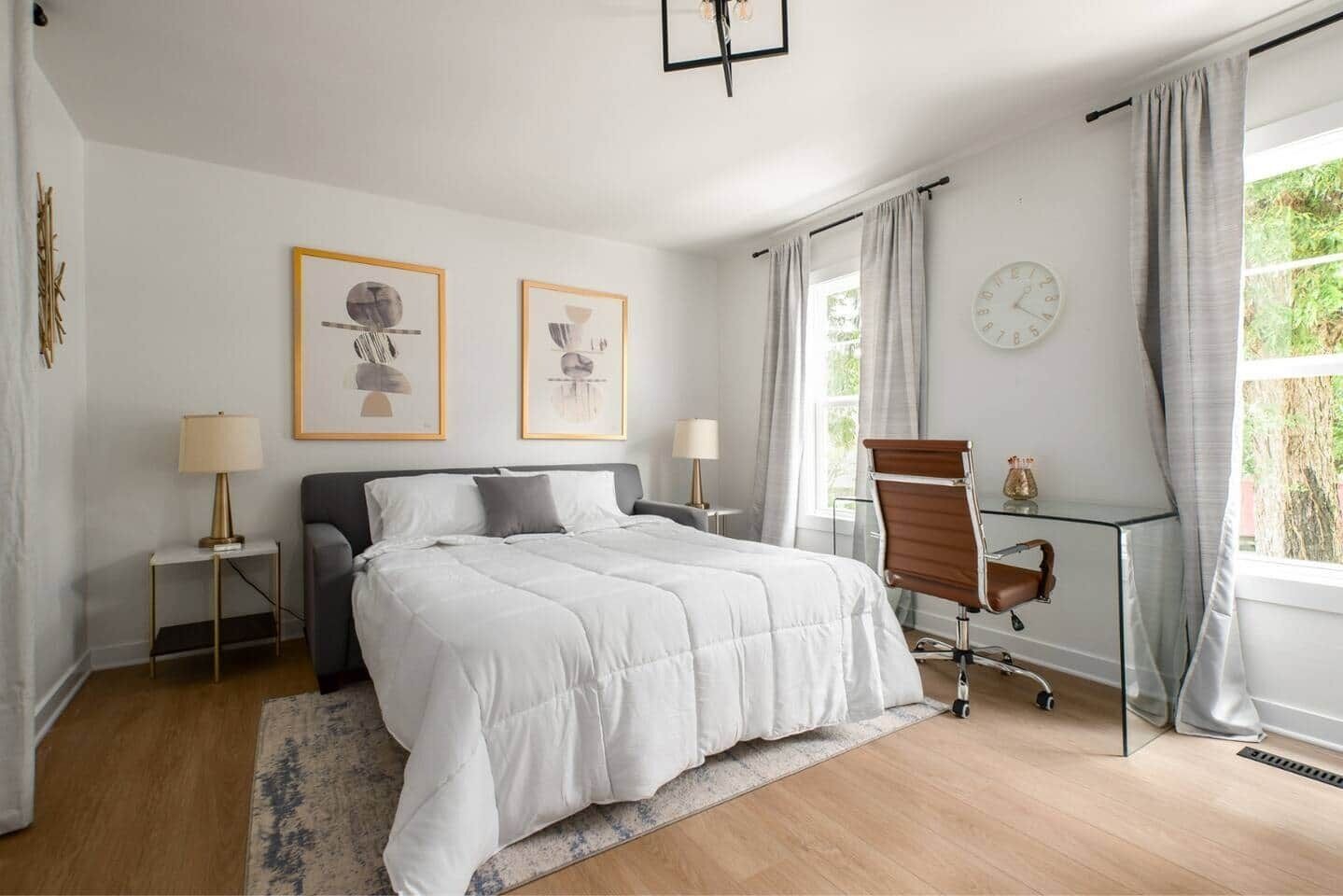 A bedroom with a bed, two bedside tables with lamps, a chair, a desk with a chair, a clock, and a window with curtains.