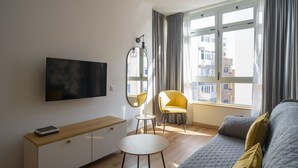A modern, well-lit room with a large window, a flat-screen TV mounted on the wall, a small round table, a yellow chair, and a grey sofa with cushions.